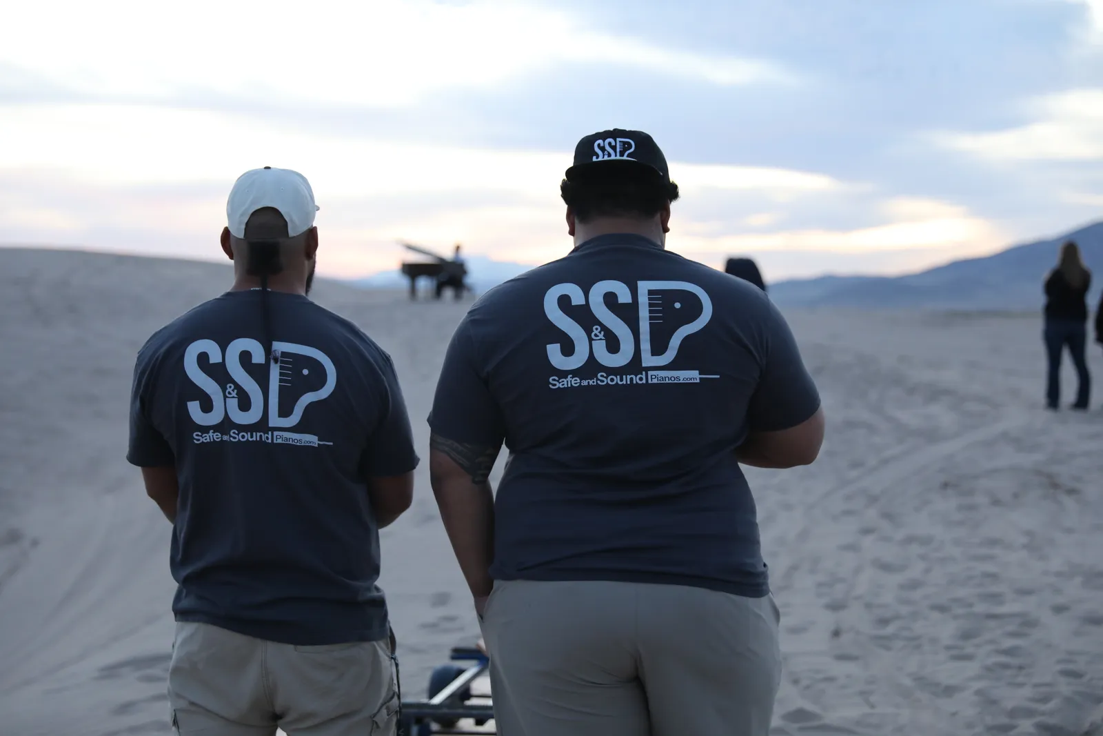 Two Safe and Sound Pianos crew members in branded shirts walking toward a grand piano staged on Little Sahara sand dunes at dusk.
