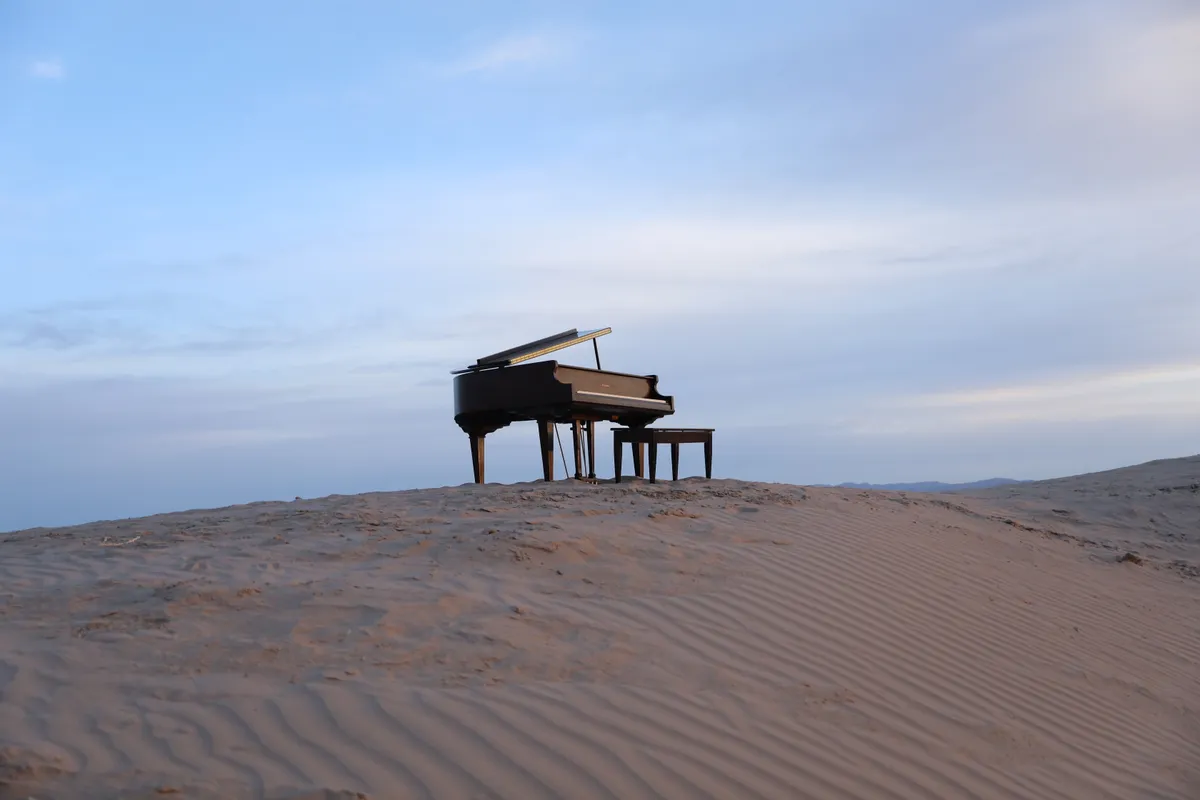 A black grand piano placed on a Little Sahara sand dune ridge against a soft sunset sky.