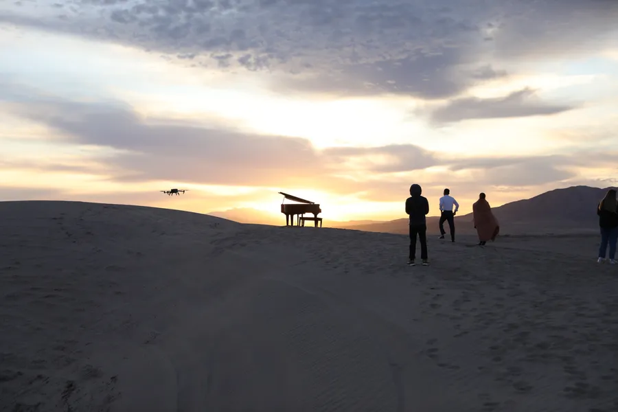 Sunset at Little Sahara: a brown grand piano glows against the horizon while a camera drone hovers and the crew looks on.