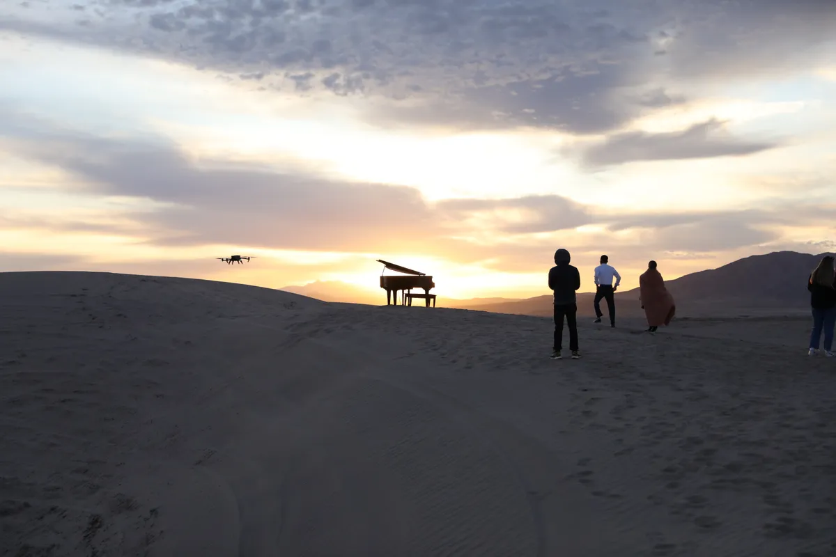 Sunset at Little Sahara: a brown grand piano glows against the horizon while a camera drone hovers and the crew looks on.