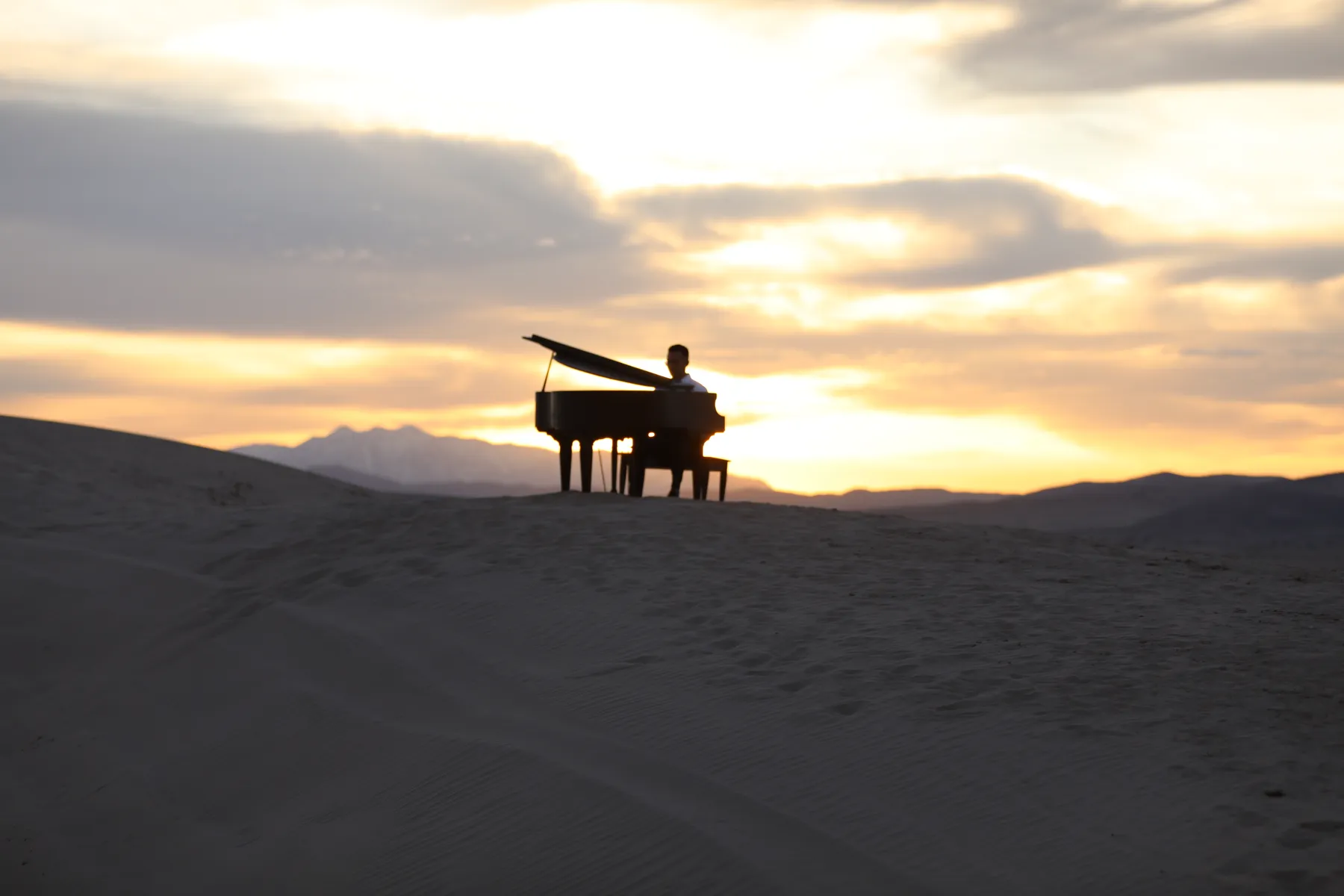 A pianist silhouetted at a grand piano on a Little Sahara dune as the sun sets behind Utah's mountains.