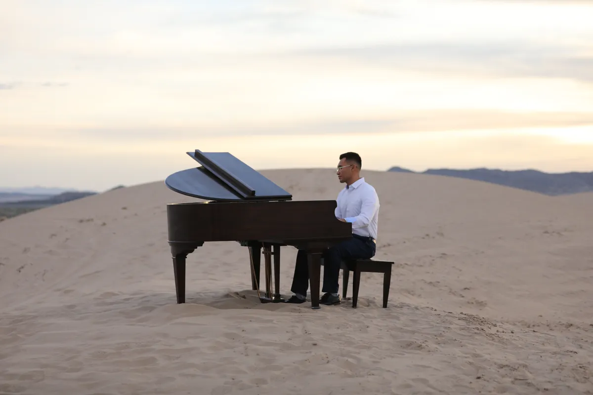 A pianist in a white shirt at a brown grand piano on a Little Sahara dune at twilight, mountains in the distance.