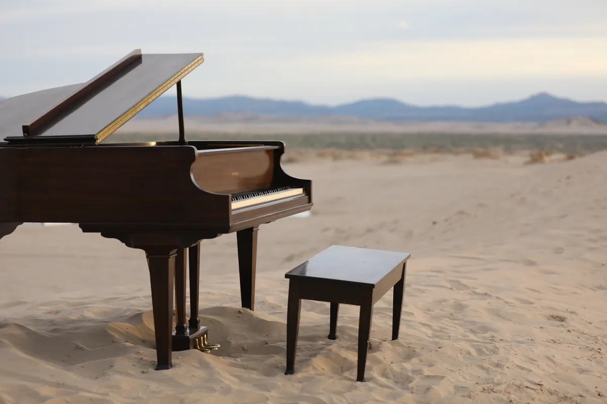 Wide view of a brown grand piano and bench alone on a sand dune at Little Sahara, mountains in the distance.