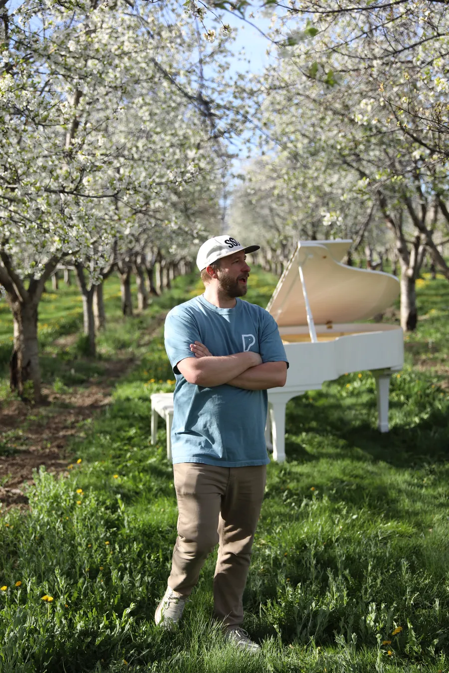 Jonny, co-owner of Safe and Sound Pianos, leaning on a white grand piano in a Santaquin cherry orchard in bloom.