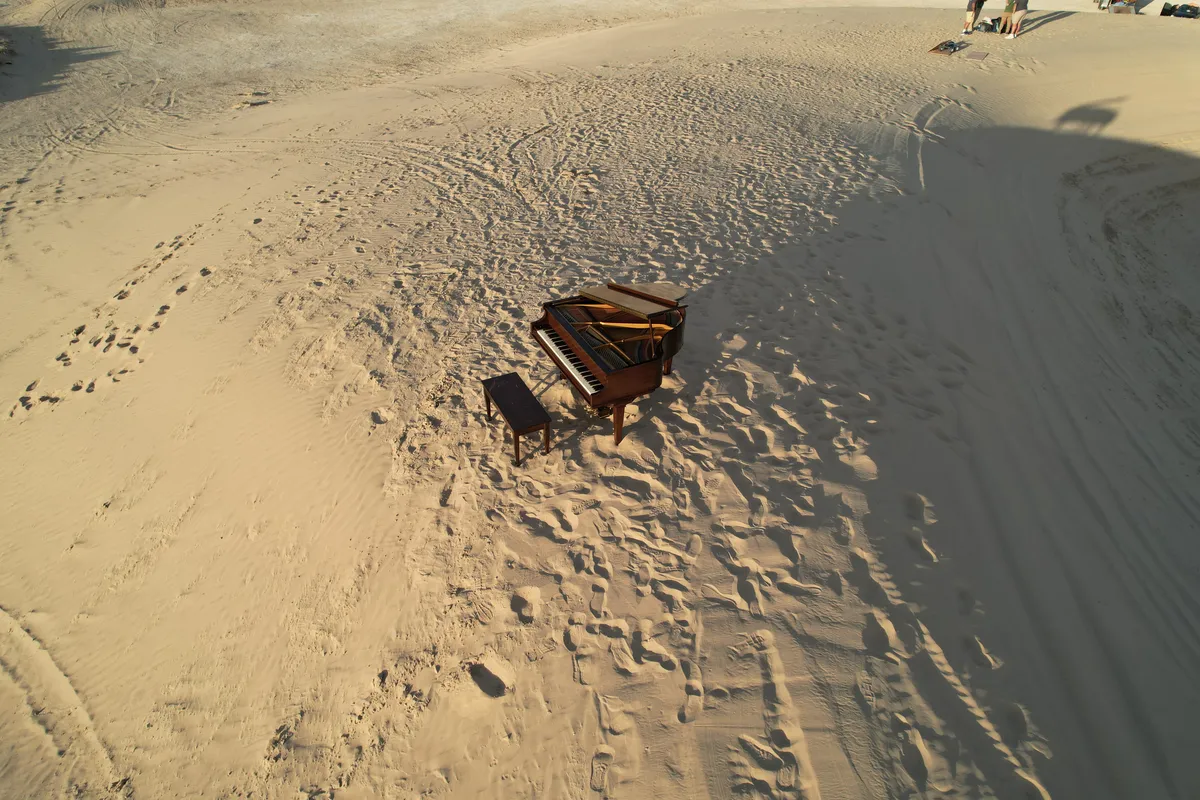 Drone aerial of Little Sahara dunes at sunset — a grand piano with a figure walking on the sand below.