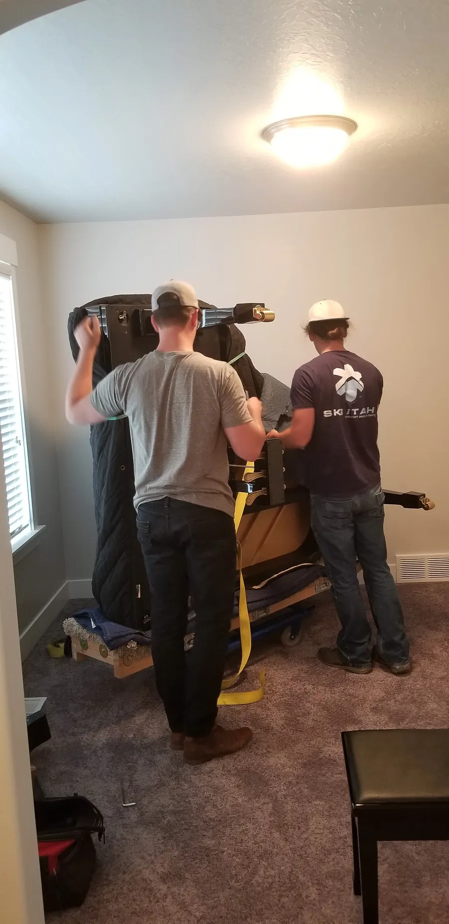 A wrapped upright piano staged on a moving dolly inside a Utah residence, crew prepping for the lift.