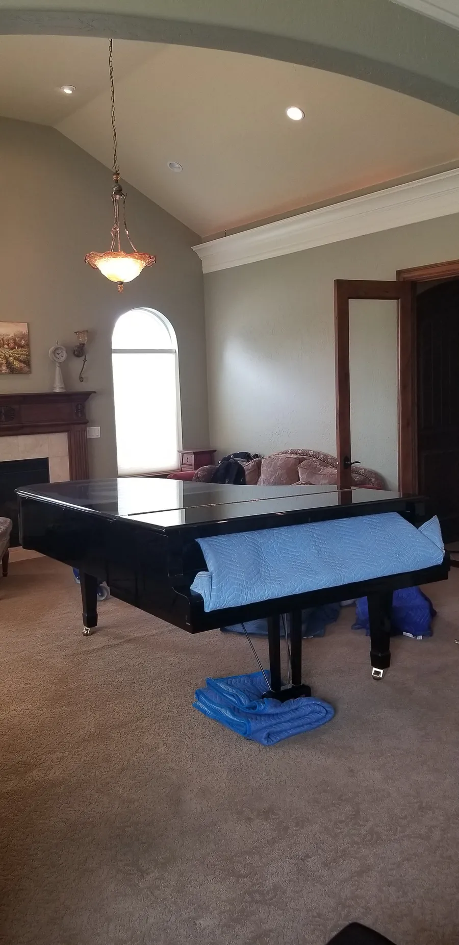 A black grand piano wrapped in moving blankets sits in a vaulted-ceiling living room, ready to be loaded out by Safe and Sound Pianos.