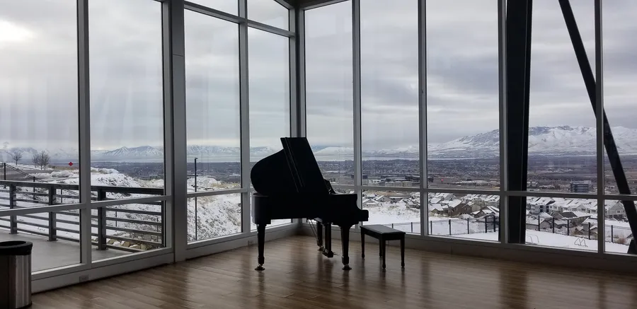 A black grand piano placed in a glass-walled room overlooking the Wasatch Mountains and the snow-covered Salt Lake Valley.