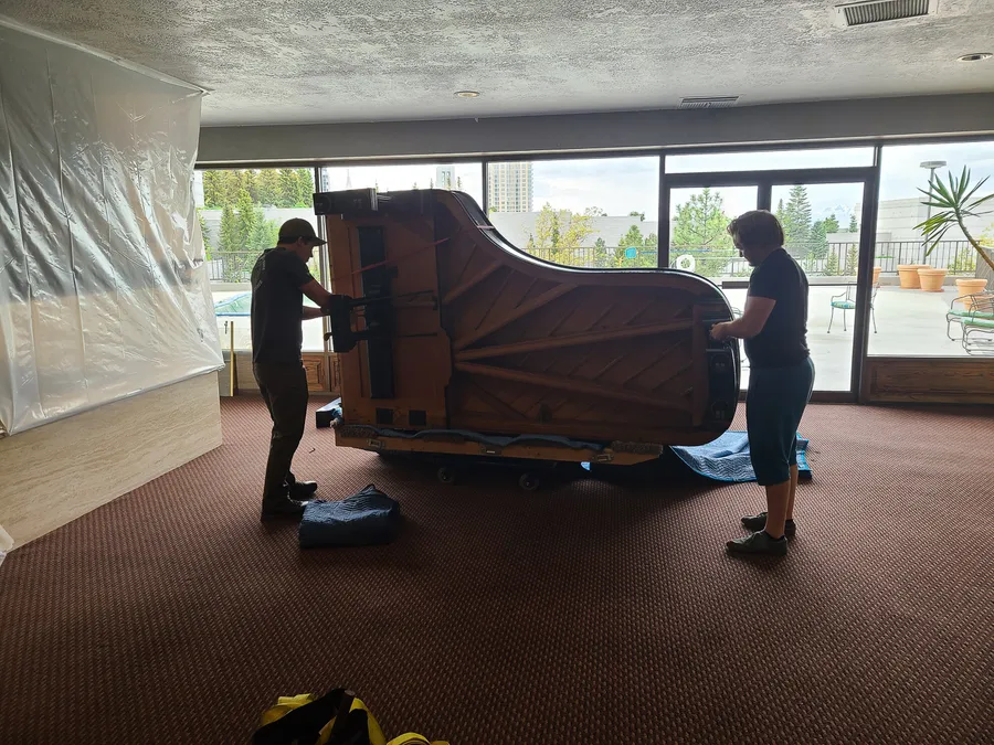 Two crew members rolling a wrapped grand piano on a piano dolly through a venue interior.