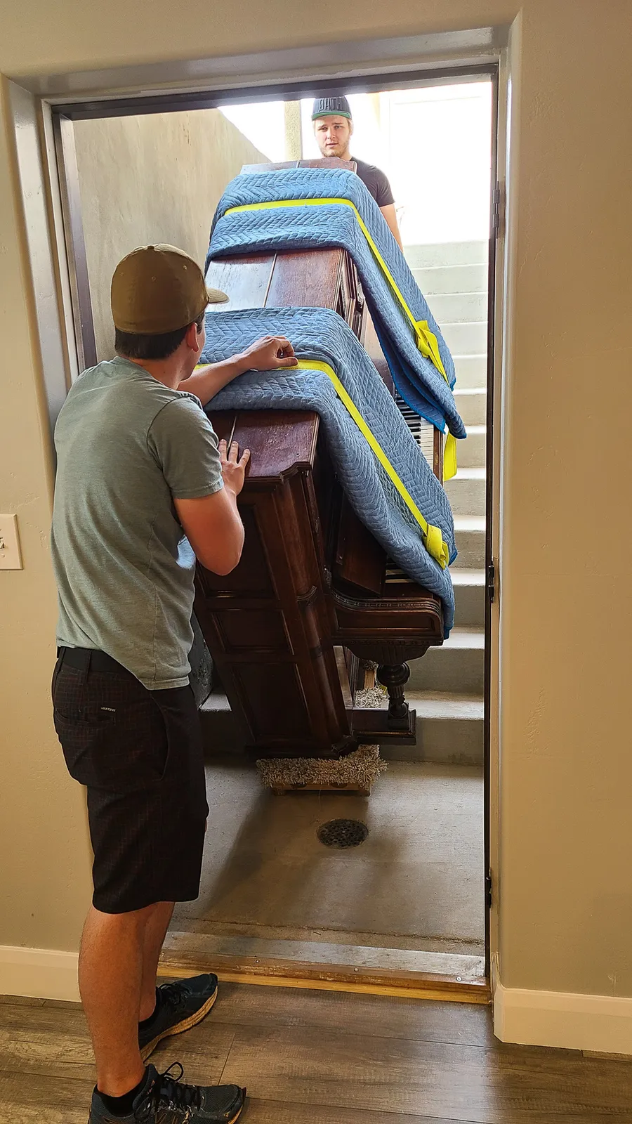Two crew members guide a wrapped upright piano through a doorway at the top of an entry-stair ramp.
