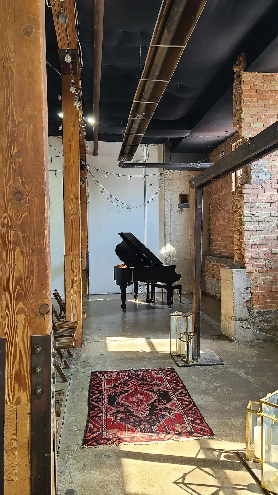 A black grand piano placed in an industrial brick venue with reclaimed beams and a Persian rug — a Safe and Sound Pianos rental delivery.